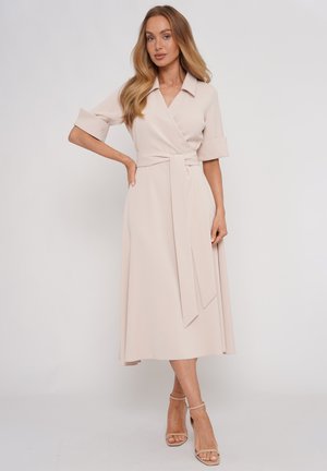 Kobieta z długimi blond włosami, ubrana w jasnobeżową midi sukienkę kopertową z podwiniętymi rękawami i szpilki z paskiem wokół kostki, stojąca na jednolitym tle.