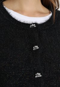 Musta pörröinen neuletakki, jossa on hohtava pinta ja kolme koristeellista hopeanväristä kukkamuotoista nappia edessä.