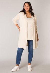 Beige open cardigan met lange mouwen, gladde textuur, gecombineerd met een bijpassend topje en blauwe slim-fit jeans, afgemaakt met witte sneakers.