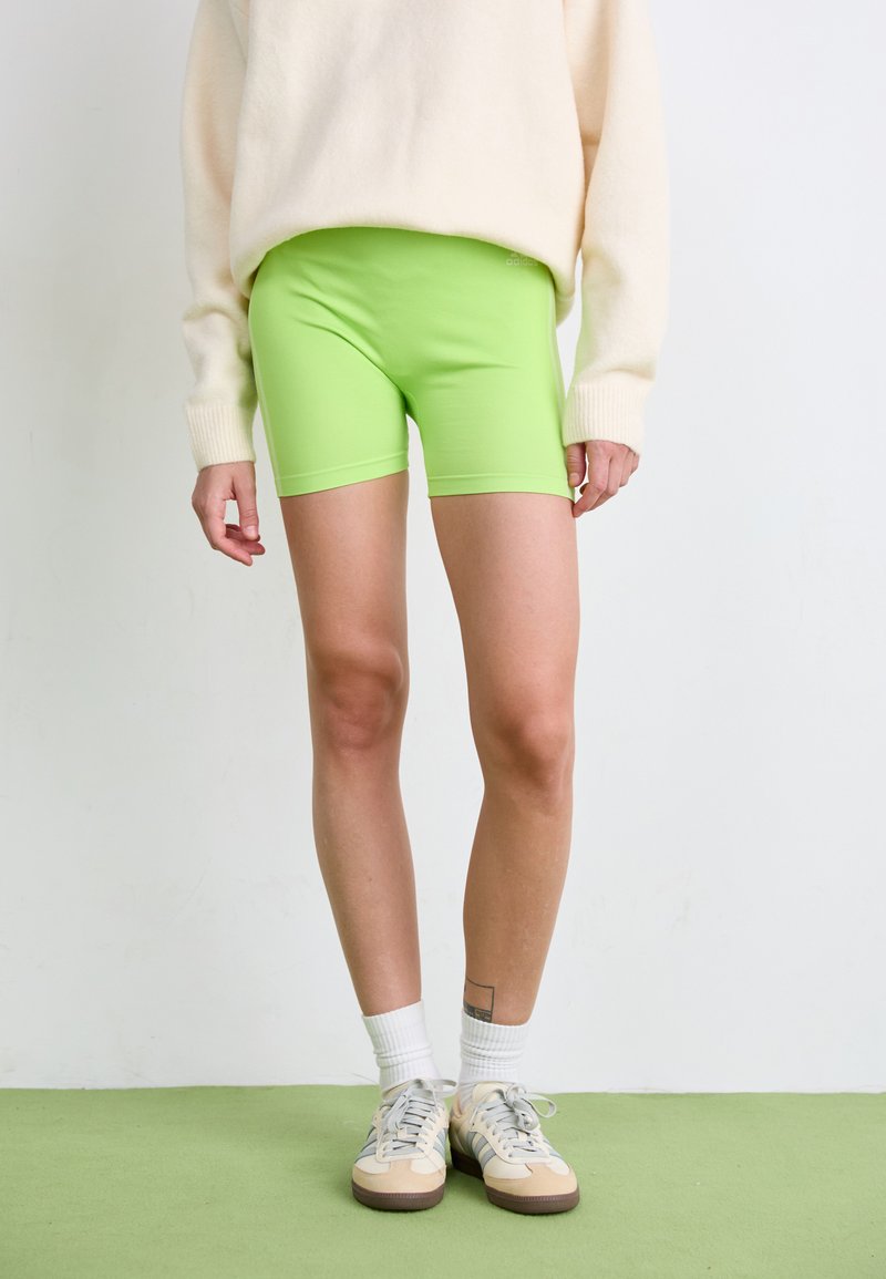 Neon groene sportshorts met een gladde textuur, gecombineerd met een witte trui en beige schoenen, tegen een bleke achtergrond.