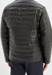 Uomo che indossa una giacca di pelle trapuntata nera, vista di spalle, abbinata a pantaloni blu scuro su uno sfondo neutro.