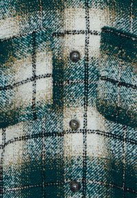 Camicia di flanella a quadri verdi e crema con cuciture nere, dotata di due tasche sul petto e bottoni grigi su un tessuto testurizzato.