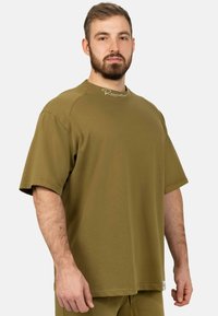 T-shirt verde oliva a maniche corte in cotone, con colletto rotondo e un sottile logo vicino al collo. Texture liscia.