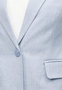 Blazer bleu clair texturé, boutonné une fois, porté par-dessus un haut blanc côtelé, montrant les détails du revers et des poches à rabat.