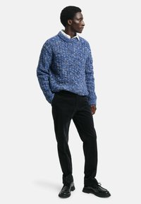 Niebieski sweter dzianinowy o teksturowanym wzorze, zestawiony z czarnymi spodniami sztruksowymi i czarnymi butami lace-up. Model stoi na neutralnym tle.