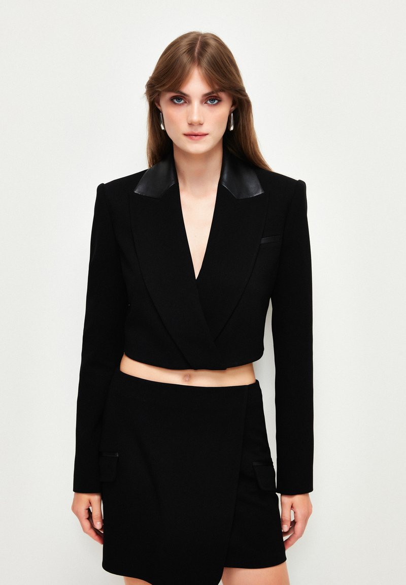 Svart kort blazer med en slät läderkrage, parat med en svart mini kjol som har sidofickor och en inslagsdesign.