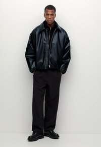Blouson aviateur en cuir noir avec un col montant, fermeture éclair et coupe décontractée. Associé à un pantalon noir et des chaussures noires volumineuses.