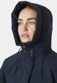 Chaqueta con capucha de color navy, con alto cuello, cuerdas ajustables y cierre frontal con cremallera. Textura suave y diseño contemporáneo.