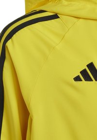 Giacca sportiva gialla con strisce nere ai lati e logo ricamato. Il materiale sembra liscio con un design leggero.
