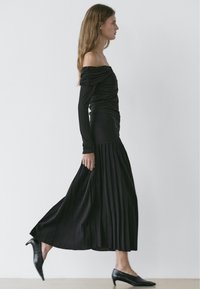 Woman walking in a black off-shoulder top and long pleated skirt wearing black low-heeled shoes against a plain background.