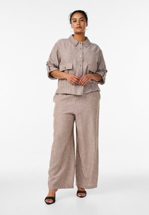 Kobieta ubrana w beżową koszulę na guziki z podwiniętymi rękawami oraz pasujące szerokie spodnie, do tego czarne sandały na obcasie na białym tle.