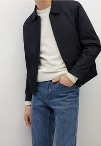 Mann in einer schwarzen Zip-Jacke über einem weißen gerippten Pullover mit blauen Jeans, eine Hand in der Tasche und die andere entspannt an der Seite.