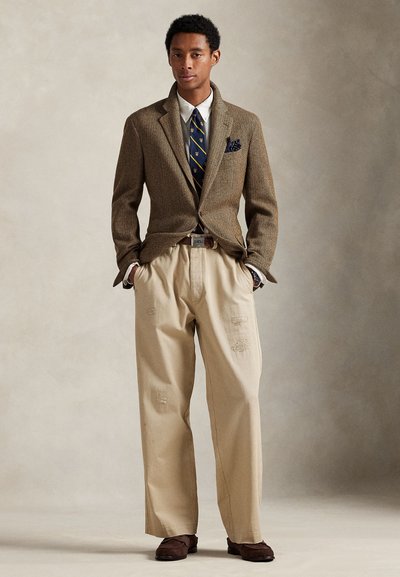 Brun blazer i fiskebensmønster, hvit skjorte, marineblått slips med gullaksenter, beige bukser med ujevne lapper, og brune sko.