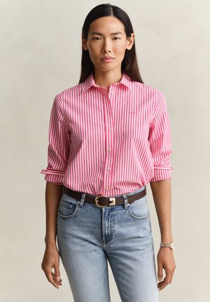 Woman wearing a pink and white vertically striped button-up shirt, light blue jeans, and a black belt with a gold buckle, standing neutral.