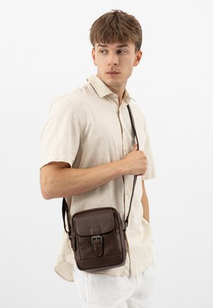 Młody mężczyzna w beżowej koszuli na krótki rękaw z zapięciem na guziki oraz białych spodniach, niosący brązową skórzaną torbę na ramię.