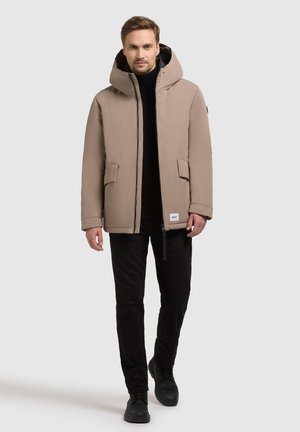 Beige vadderad jacka med stor huva, framdragkedja, två sidofickor och en logotypficka vid fållen. Bärs över en svart tröja och byxor.