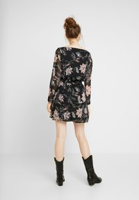 Černé květinové šaty s dlouhými průhlednými rukávy, které mají růžové a modré květinové vzory. Šaty se stahují v pase, doplněné o černé boty.