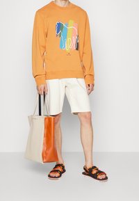 Sudadera naranja con un gráfico de caballo, pantalones cortos crema, sandalias negras con acentos naranjas y una bolsa de mano beige y naranja.