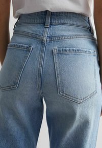 Vaqueros de mezclilla azul claro con cintura alta y bolsillos traseros, llevados por una persona con una camiseta blanca, mostrados desde atrás a la altura de las caderas.