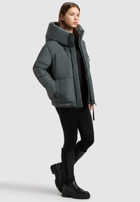 Kvinde iført en mørkegrå pufferjakke med høj krave, sorte leggings og sorte knæhøje støvler, der står mod en ensfarvet lys baggrund.