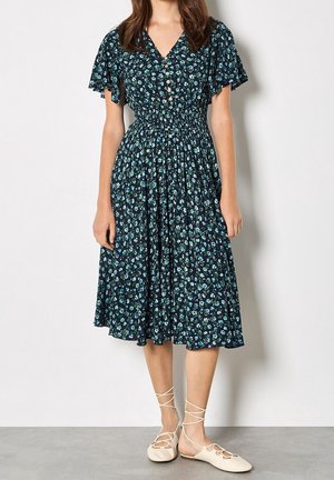 Femme portant une robe midi fleurie bleu foncé avec de courtes manches volantes, une taille froncée, des boutons à l'avant et des chaussures plates blanches à lacets.