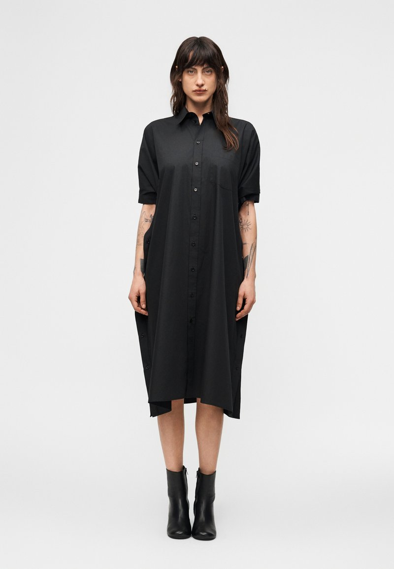 Γυναίκα με τατουάζ που φορά μαύρο midi φόρεμα με κουμπιά και μαύρα μποτάκια μέχρι τον αστράγαλο, στέκεται μπροστά σε απλό φωτεινό φόντο.