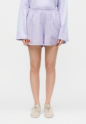 Person iført lavendel ternede shorts med elastisk linning, en lavendel langærmet top og beige sneakers, stående mod en ensfarvet baggrund.