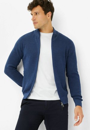 Marineblaue, durchgehend zu schließende Strickjacke aus strukturiertem Strickstoff. Mit Stehkragen und gerippten Bündchen, getragen über einem weißen T-Shirt.