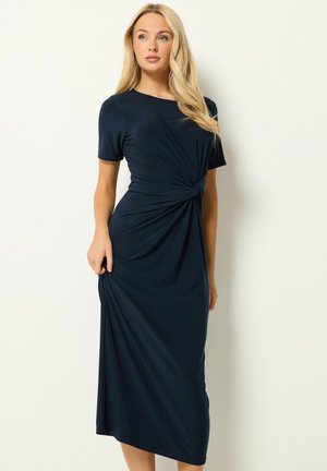 Femme aux longs cheveux blonds portant une robe midi bleu marine à manches courtes avec un détail nœud torsadé à la taille.