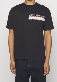 Persona que lleva una camiseta negra de Calvin Klein Jeans con texto blanco y rosa en el pecho y jeans oscuros.