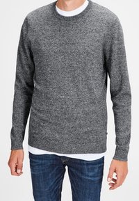 Grå stickad tröja med rund halsringning och långa ärmar, i kombination med ett vitt innerskikt. Tyget har en slät yta och en lätt mönstring.