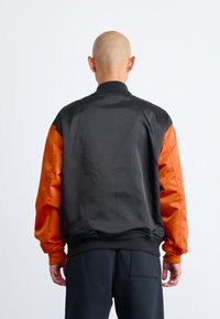 Person with a shaved head wearing a black and orange bomber jacket facing away against a plain white background.