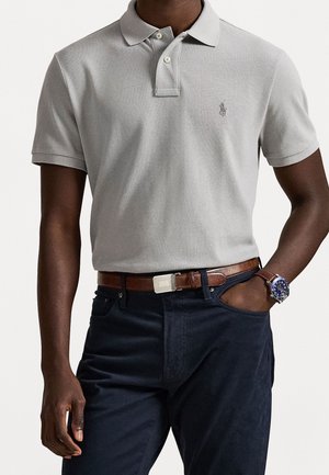 Mann trägt ein hellgraues Poloshirt mit kurzen Ärmeln, dunkle Hose, braunen Ledergürtel und eine Uhr mit blauem Zifferblatt am linken Handgelenk.