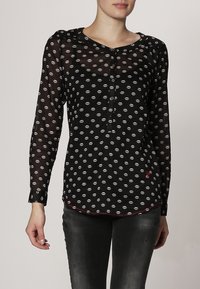 Blouse noire transparente avec imprimé de lèvres blanches, manches longues, patte de boutonnage, et accent rouge à l'ourlet. Texture douce avec un design léger.