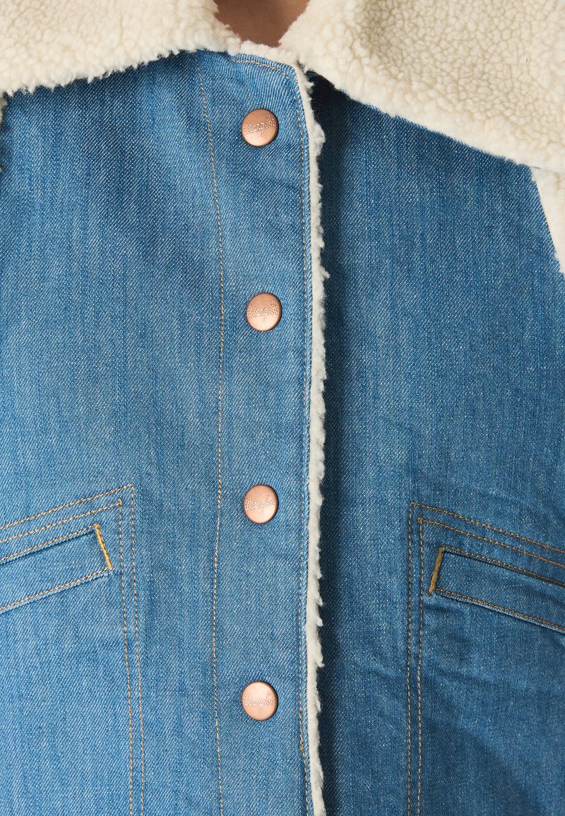 Jeansowa kurtka z kołnierzem z owczej wełny, z mosiężnymi guzikami, bocznymi kieszeniami i kontrastowym przeszyciem na niebieskim materiale. Teksturowane wykończenie.