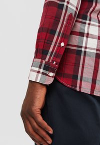 Braccio sinistro con camicia a quadri rossa, bianca e nera a maniche lunghe con bottoni bianchi, appoggiato accanto a pantaloni blu scuro.