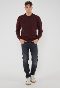 Giovane uomo in piedi con le mani in tasca, indossa un maglione bordeaux, jeans scuri e sneaker bianche su uno sfondo semplice.