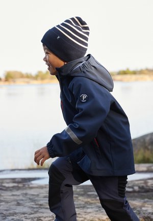 Marineblauwe waterdichte jas met capuchon, reflecterende accenten en ritszakken; gecombineerd met donkere broek en een gestreepte gebreide beanie.