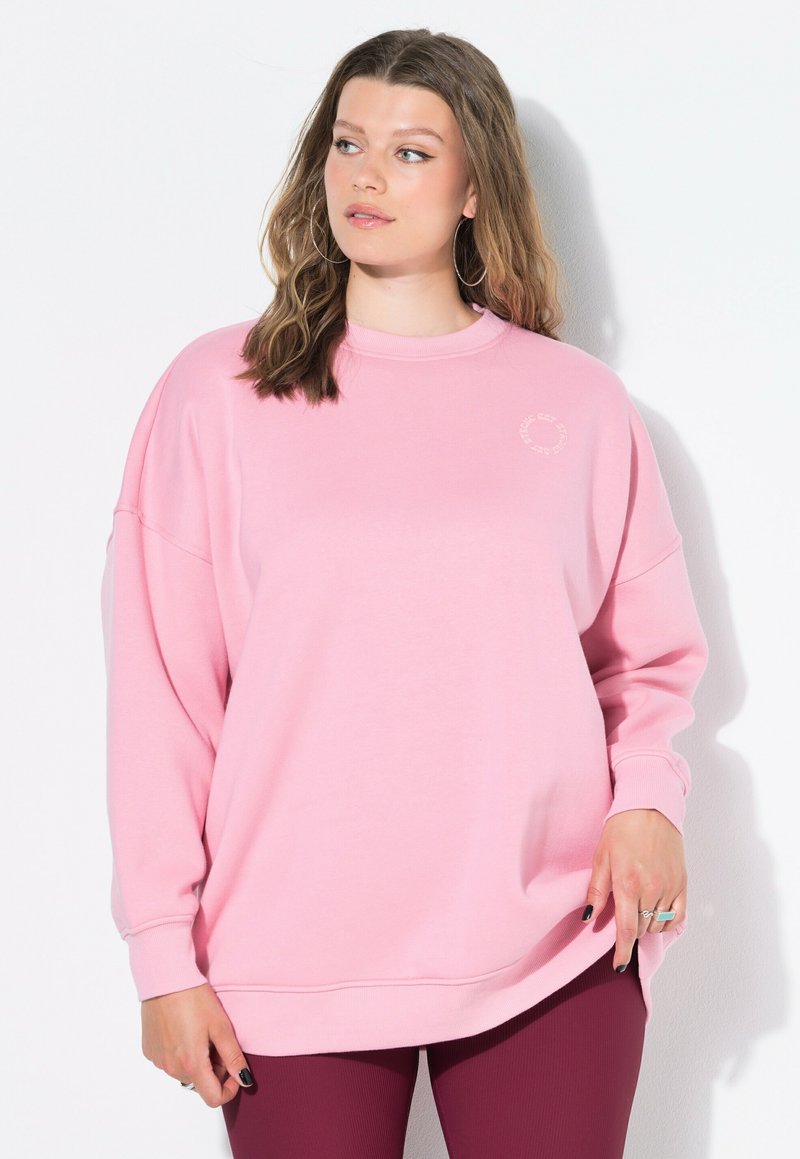 Sudadera de gran tamaño en rosa claro con cuello redondo y mangas largas; presenta un pequeño logotipo en el pecho y puños y dobladillo acanalados.