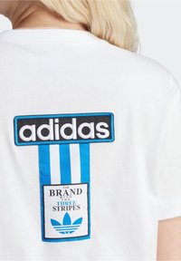 Vit bomull t-shirt med en svart och blå "adidas" logopatch och en blå randig design nedanför, där det står "THE BRAND WITH THREE STRIPES."