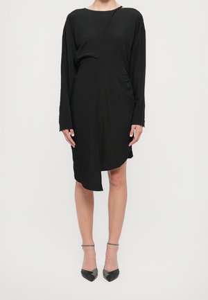 Femme portant une robe asymétrique noire à manches longues avec des talons pointus noirs à bride cheville sur un fond blanc uni.