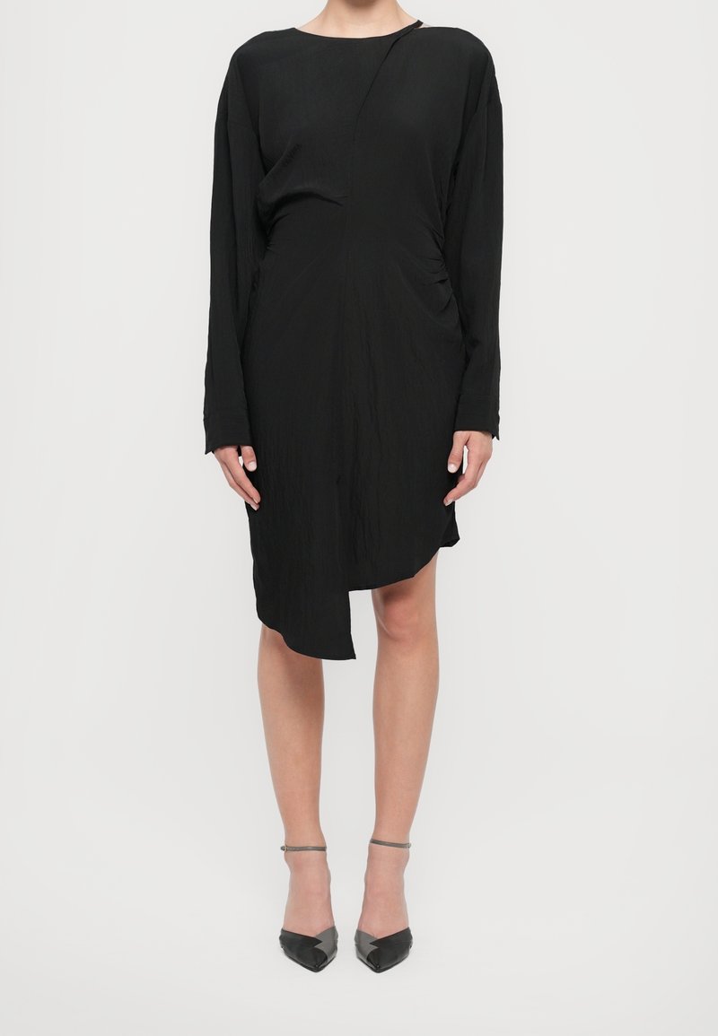 Femme portant une robe asymétrique noire à manches longues avec des talons pointus noirs à bride cheville sur un fond blanc uni.