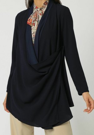 Blusa drapeada en navy con mangas largas, detalle de plisado frontal y una textura suave, superpuesta sobre una camisa con cuello estampado.