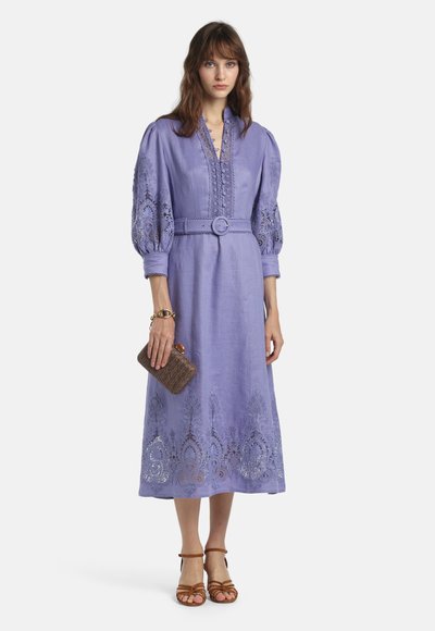 Frau trägt ein lila Midikleid mit Gürtel und puffigen Spitzenärmeln, hält eine braune Clutch und trägt braune Riemchensandalen.