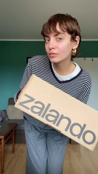 Brązowy kartonowy pudełko z napisem "zalando" w szarym kolorze. Osoba jest ubrana w pasiaste czarno-białe sweter i szare szerokie spodnie.