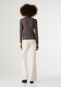 Kortsukel on lokkis juustega inimene, kes kannab pika varrukaga triibulist särki ja kõrge talje ägedaid valgeid teksaseid koos mustade kontsakingadega, seistes ühetaolise tausta ees.