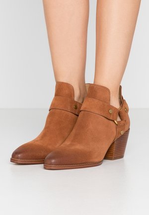 Paire de bottines en daim marron avec talons épais superposés et détails de sangles, portées sur jambes nues devant un fond uni.