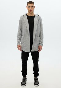 Joven de pie, vistiendo un cárdigan con capucha de abertura frontal en gris claro, camiseta negra, pantalones cargo negros y zapatillas en gris y negro sobre un fondo blanco.