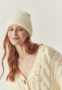 Beanie de tricot de culoare crem deschis, cu textură în bucle. Cardigan tricotat gros, cu un model de cablu și decolteu adânc în formă de V. Materiale moi și confortabile.