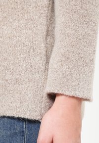 Beiger Strickpullover mit strukturierter Oberfläche. Verfügt über ein kurzes Design und weite Ärmel, kombiniert mit blauen Jeans.